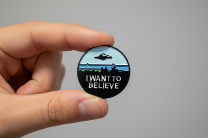 A circular enamel pin held in a hand, featuring a black UFO flying over a landscape with the text 'I WANT TO BELIEVE' in silver lettering.