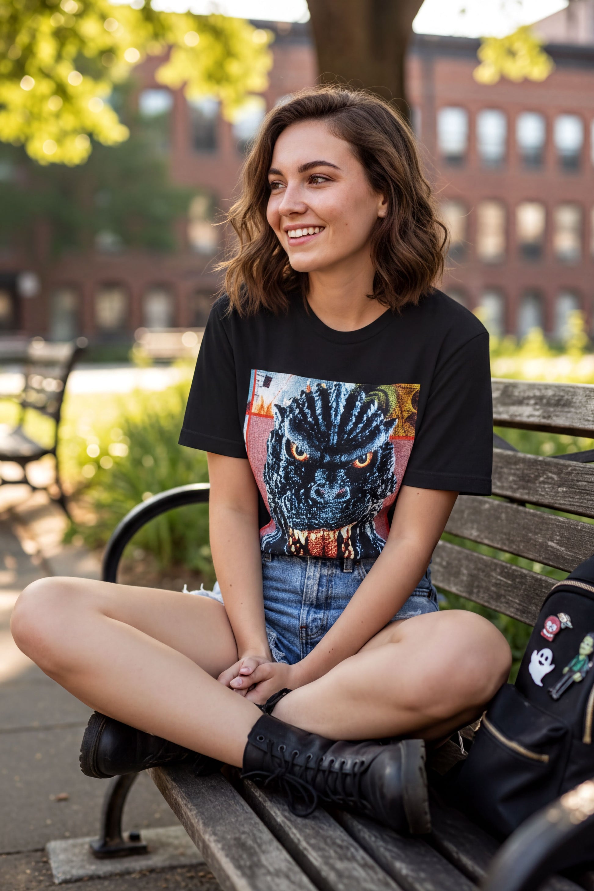 A young woman is sitting on a wooden bench in a park, wearing a black t-shirt with a graphic design and blue shorts.