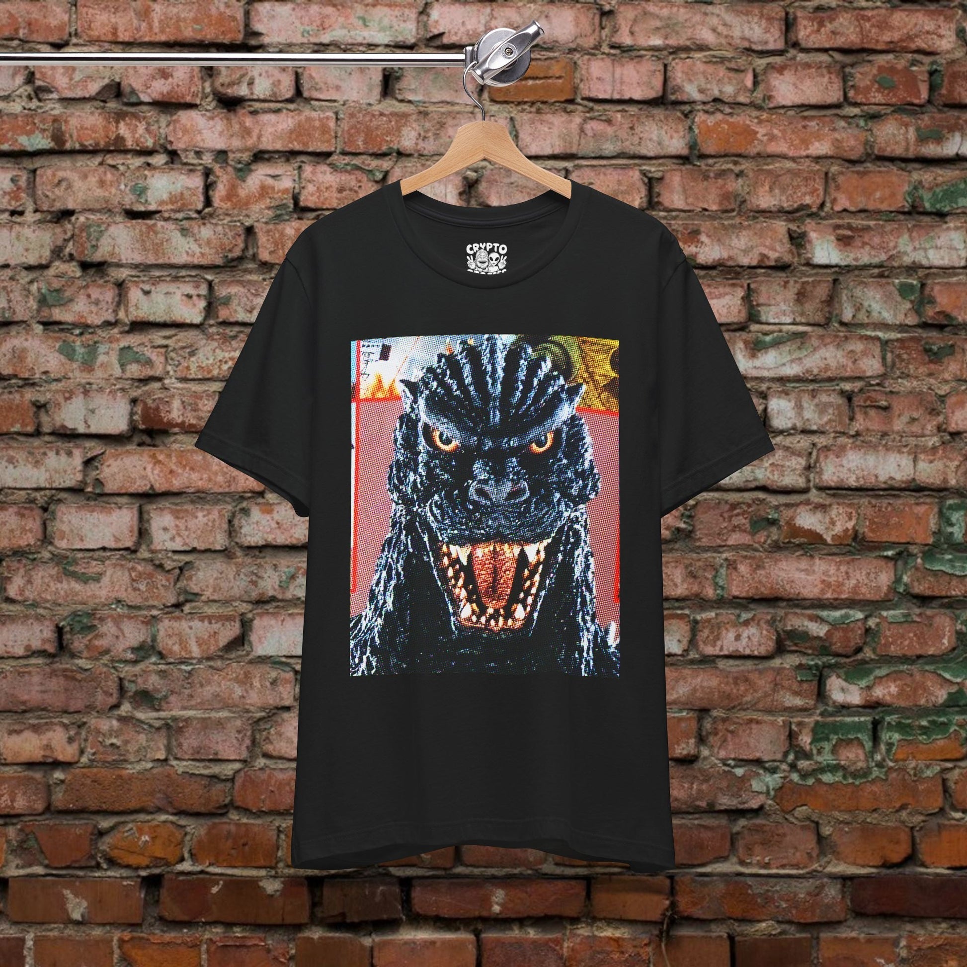 A black t-shirt with a graphic of a large, menacing-looking monster with a fierce expression and open mouth, hanging on a wooden hanger against a brick wall.